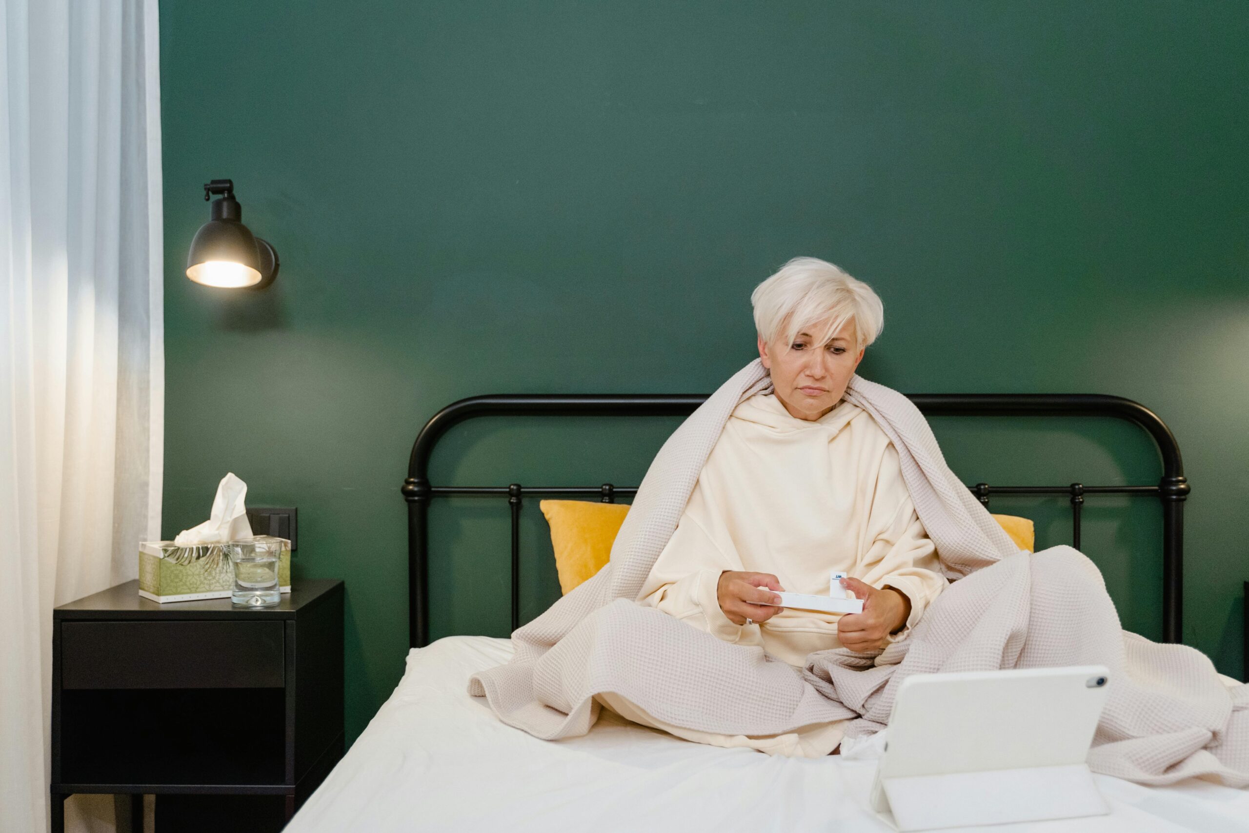 Senior woman in bed using a tablet for telemedicine during illness, wrapped in blanket.